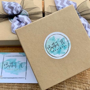 Puede incluir: Dos cajas de cart&oacute;n marr&oacute;n con cinta de chevron gris y blanco y tul negro. Las cajas tienen una pegatina con el texto "Button It Designs." en ellas. Una tarjeta de visita con el mismo logotipo est&aacute; en primer plano.