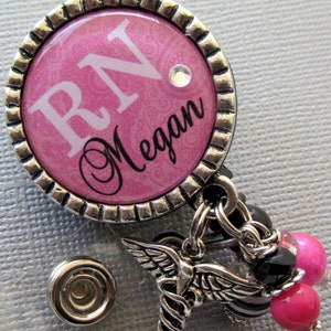 May include: A pink and silver badge holder with a white 'RN' and the name 'Megan' in black script. The badge holder has a silver charm with a caduceus and wings.