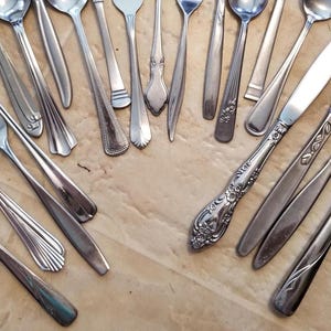 May include: An assortment of vintage silverware, including forks, spoons, and knives, arranged on a light brown surface. The silverware features various handle designs, from simple to ornate, showcasing a range of styles and patterns. The utensils are made of a shiny, metallic material.