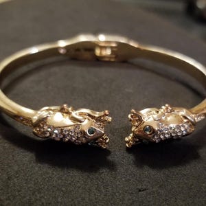 May include: Gold-toned hinged bracelet featuring two ornate mouse heads encrusted with clear crystals and small blue gemstones for eyes. The bracelet is open, showcasing the clasp mechanism. The overall design is detailed and elegant.