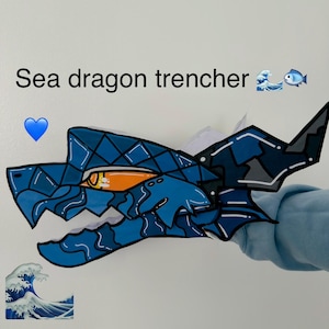 May include: A blue paper craft sea dragon head with a geometric pattern, a sharp orange eye, and black outlines. The text "Sea dragon trencher" is printed above the dragon head, accompanied by a blue heart, a wave, and a fish emoji.
