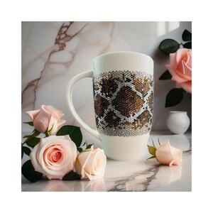 Könnte beinhalten: Weiße Keramiktasse mit einem Schlangenhautmuster aus Strasssteinen. Die Tasse ist von rosa Rosen umgeben.