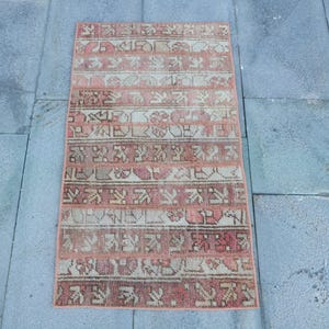 May include: Rectangular, vintage-style rug with a faded, distressed appearance. The rug features a repeating pattern of geometric shapes and floral motifs in shades of pink, beige, and brown. The rug is likely made of wool or a similar natural fiber.