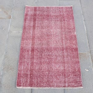 May include: A rectangular area rug featuring a faded, distressed texture in shades of deep crimson and muted rose. The rug displays a subtle, worn-in pattern with vertical striations and a short, fringed edge along the top and bottom borders.