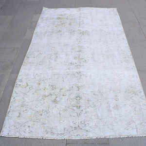 May include: A rectangular, off-white rug with a faded, floral pattern. The rug has a distressed look with muted green and brown accents. The rug is placed on a gray stone surface, showcasing its size and design.