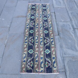 Puede incluir: Una alfombra rectangular de estilo vintage con fondo azul oscuro y rayas verticales. Las rayas presentan patrones repetidos en tonos verdes, rosas y azules. La alfombra está colocada sobre una superficie de piedra gris.