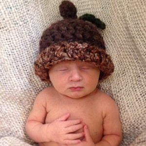 May include: A newborn baby wearing a brown and white crocheted acorn hat with a green leaf detail. The baby is lying on a white blanket and has their eyes closed.