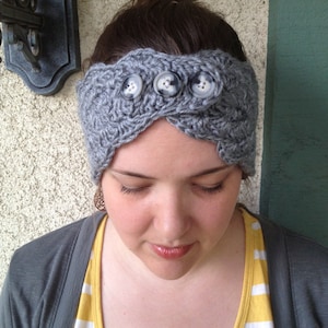 Puede incluir: Una diadema de punto gris con tres botones grandes. La diadema la lleva puesta una persona con el pelo castaño, recogido en un moño suelto.