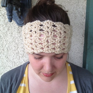 May include: A close-up of a woman wearing a crocheted headband in a cream color. The headband has a square pattern and is worn over her brown hair. She is wearing a gray cardigan over a yellow and white striped shirt.