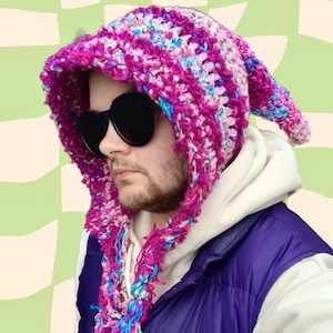 May include: A pink, blue, and white crocheted hooded scarf with a long, flowing tail. The scarf is worn by a person wearing a white hoodie and a purple vest.