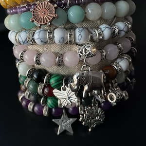 May include: A stack of bracelets with various charms, including a butterfly, an elephant, a star, and a tree. The bracelets are made of different types of beads, including amethyst, rose quartz, and agate.