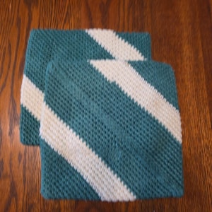 Peut inclure: Deux carrés tricotés à rayures turquoise et blanches sont présentés sur une surface en bois. Les carrés ont une rayure blanche diagonale sur le fond turquoise. La texture du tricot est visible, et les carrés sont bien disposés.