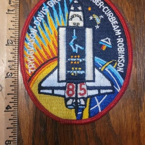 Puede incluir: Parche bordado circular con un diseño de transbordador espacial y el número 85. El parche tiene un borde rojo e incluye texto como "DAVIS BROWN ROMINGER CURBEAM ROBINSON". El fondo tiene un diseño colorido.
