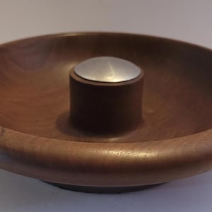 Puede incluir: Un cuenco de madera con un poste cilíndrico central coronado por una tapa de metal plateado. El cuenco es de color marrón cálido y tiene un borde liso y redondeado. El diseño es simple y elegante.