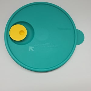 以下が含まれることがあります： 黄色の丸いボタンと小さなハンドルが付いた、ターコイズ色の Tupperware の蓋。蓋には Tupperware のロゴとテキストがあります。蓋は丸く、縁が湾曲しています。
