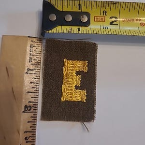 May include: A small, olive green patch with a gold embroidered letter "E". The patch is rectangular and measures approximately 1.25 inches in length. A yellow measuring tape is in the background.