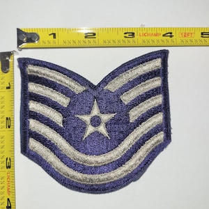 Peut inclure: Écusson militaire brodé bleu et argenté. L'écusson présente une étoile blanche centrale et plusieurs rayures argentées sur fond bleu. L'écusson mesure environ 10 cm de large.