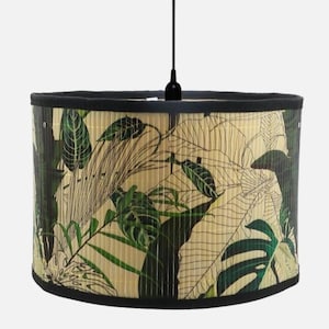 May include: A cylindrical pendant lamp shade featuring a bamboo-like texture with a printed pattern of green tropical leaves on a light beige background. The shade is framed by a solid black trim along the top and bottom edges and hangs from a cord.