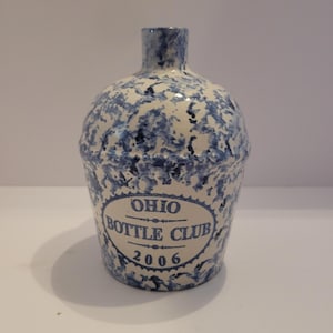 Puede incluir: Botella de cerámica con un patrón moteado azul y blanco. La botella tiene un cuerpo redondeado y un cuello estrecho. Las palabras "OHIO BOTTLE CLUB 2006" están impresas en la parte delantera en un óvalo azul.