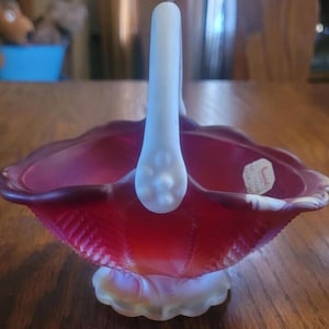 May include: A ruby red glass basket with a white handle. The basket has a scalloped rim and a textured design on the sides. The handle is smooth and curved. The basket sits on a white, petal-shaped base.