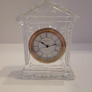 Puede incluir: Un reloj de cristal de Waterford con una esfera con borde dorado y números romanos. El reloj está alojado en una estructura de cristal transparente que se asemeja a un pequeño edificio con una parte superior decorativa.