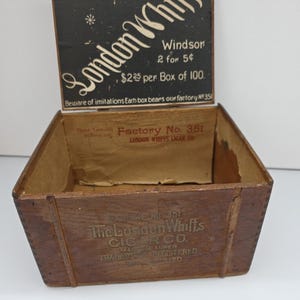 Puede incluir: Una caja de cigarros de madera antigua y abierta. La tapa es negra con texto blanco que dice "London Whiffs" y "Windsor". La caja es marrón con el texto "Factory No. 351" y "The London Whiffs Cigar Co."