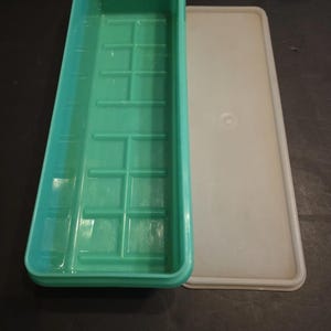 May include: A rectangular, teal-colored plastic container with a grid pattern inside, next to a white lid. The container is open, revealing its interior. The lid is rectangular and has a slightly raised edge.