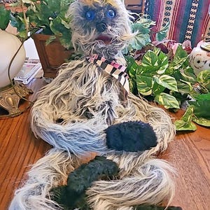 May include: A handmade, whimsical plush creature with a shaggy, gray and white fur body. It has a colorful, rainbow-hued headpiece, blue eyes, and a striped scarf. The plush is designed to resemble a fantasy character.