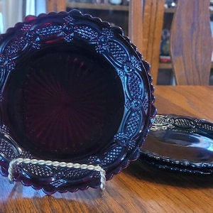 Könnte beinhalten: Ein Satz tiefroter Glasplatten mit einem dekorativen geprägten Muster. Die Teller haben einen gewellten Rand und werden auf einem silberfarbenen Metallständer präsentiert. Die Teller stehen auf einem Holztisch.