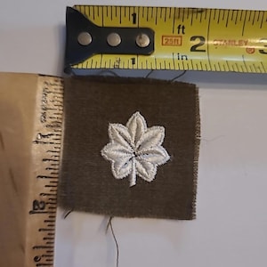 May include: A military insignia patch featuring a white embroidered leaf design on a dark green square. A yellow and black measuring tape is positioned above the patch, showing measurements in inches.