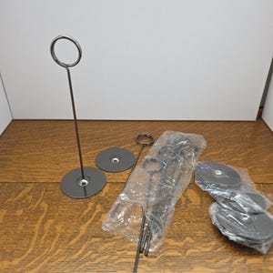 Puede incluir: Una colección de portatarjetas de color plateado y bases redondas. Algunos soportes están empaquetados en plástico transparente. Los soportes tienen una parte superior circular para sujetar tarjetas. Las bases son circulares y planas.