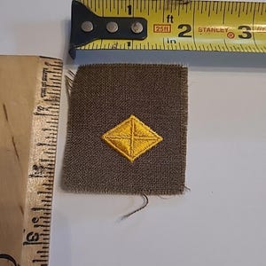 Puede incluir: Un parche de tela marrón con un diseño bordado en forma de diamante amarillo. Una cinta métrica amarilla y negra está al lado del parche. El parche mide aproximadamente 2,5 cm cuadrados.