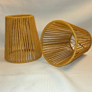 May include: Two conical lampshades constructed from mustard yellow cord wrapped around a wire frame. The shades feature a vertical, open-weave design that allows light to pass through the gaps between the individual strands of cord.