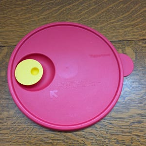 May include: A round, red Tupperware lid with a yellow insert and a small handle. The lid has a circular design with a recessed area for the insert and a small tab on the side. The lid is made of durable plastic.