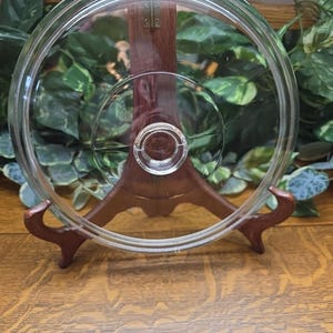 Pode incluir: Uma tampa de vidro transparente com design circular, apoiada em um suporte de madeira. A tampa tem um botão central e uma borda elevada. O suporte é marrom e tem um design curvo decorativo. O fundo apresenta folhagem verde.