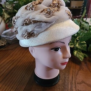 Puede incluir: Un sombrero color crema con una corona redondeada y un ala corta. El sombrero está adornado con adornos dorados y una capa de red blanca. El sombrero se exhibe en una cabeza de maniquí.