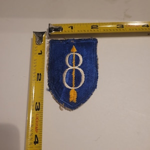 Op de afbeelding: Een blauw en wit geborduurd embleem in schildvorm. De patch heeft een wit cijfer 8 met een gele pijl erdoorheen. De patch is ongeveer 5 cm breed en 7,6 cm hoog.