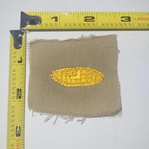 以下が含まれることがあります： 金色の刺繍デザインが施されたベージュ色の布パッチ。パッチの幅は約4.4cm、高さは2.5cmです。 金色の刺繍は、詳細で様式化されたデザインが特徴です。 パッチにはほつれたエッジがあります。