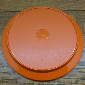Könnte beinhalten: Ein orangefarbener Tupperware-Deckel, kreisförmig, mit erhöhtem Rand und leicht strukturierter Oberfläche. In der Mitte des Deckels befindet sich das Tupperware-Logo. Der Deckel ist für einen Behälter bestimmt und besteht aus strapazierfähigem Kunststoff.