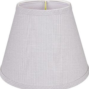 May include: A light gray, tapered drum lampshade with a visible woven fabric texture. The shade features a clean, structured silhouette with a finished top and bottom edge, and a metallic gold-toned metal fitter visible at the top opening.