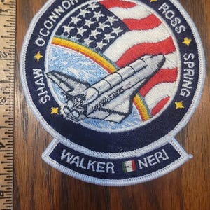 Puede incluir: Parche bordado con un diseño de transbordador espacial, una bandera estadounidense y un arcoíris. El parche incluye los nombres O'Connor, Cleave, Ross, Spring, Shaw, Walker y Neri. También es visible el texto "United States".