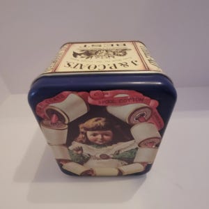 Puede incluir: Lata azul vintage con un diseño decorativo que presenta a una niña rodeada de carretes de hilo. La lata está etiquetada como "J & P Coats" y "6 Spool Cotton". La parte superior de la lata dice "BEST".