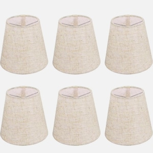 May include: Six beige fabric lampshades. Each lampshade has a conical shape with a slightly flared base and a white interior. The lampshades are arranged in two rows of three against a neutral background.