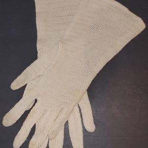May include: A pair of off-white, vintage gloves. The gloves are made of a delicate, open-weave fabric with a subtle chevron pattern. The gloves are long, extending past the wrist, and appear to be in good condition.