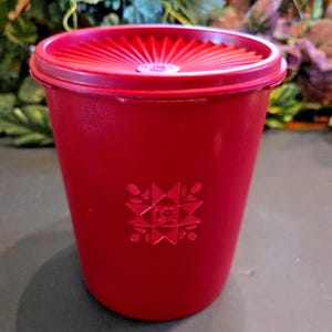 Op de afbeelding: Een rode plastic container met een bijpassende deksel. De deksel heeft een geribbeld ontwerp en de container heeft een reliëf bloemenpatroon. De container is cilindrisch en lijkt een voedselopslagartikel te zijn.