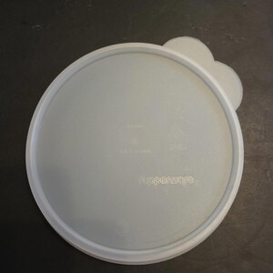 May include: A light grey Tupperware lid with a white rim and a cloud-shaped handle. The Tupperware logo is visible on the lid's surface. The lid is circular and appears to be made of plastic.