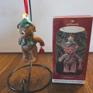 May include: A Hallmark Keepsake ornament featuring a brown bear wearing a green hat and vest, holding a red ornament. The ornament is displayed on a black metal stand. The box reads "Gift Bearers" and "NEW COLLECTOR'S SERIES."