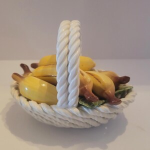 May include: A ceramic basket filled with yellow bananas. The basket has a white, twisted handle and a woven design. The bananas have brown tips and are arranged on green leaves.