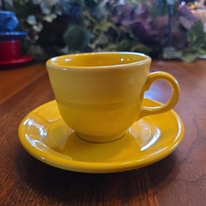 May include: A bright yellow teacup and saucer set. The cup has a rounded shape with a handle, and the saucer is circular. The set is made of ceramic and sits on a wooden surface. The background is blurred, with hints of other objects.