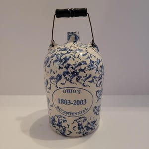 Puede incluir: Una jarra de cerámica con un patrón moteado blanco y azul. Tiene un asa negra y un asa de alambre de metal. La jarra presenta el texto "Ohio's 1803-2003 Bicentennial" en un óvalo azul. Es un objeto decorativo.